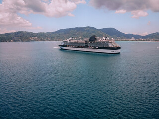 Big beautiful cruise liner sails across the picturesque bay, vast blue sea around, coastal town on the background; aerial side view.