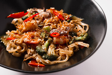 Noodles with shrimps, octopus tentacles, broccoli, chili peppers and sesame seeds in a black plate on a white background