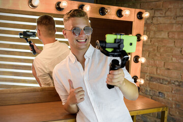 Young guy filming a blog on a mobile device using an electronic image stabilizer at the studio against the background of a mirror with illumination. Blond European man in sunglasses shoots a video