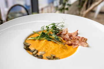 Creamy pumpkin soup with toasted bacon pieces garnished with pumpkin seeds. Healthy low-calorie soup in a white plate with a round notch