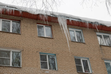 Big icicles on the roof of a townhouse on a snowy winter day among thaw. Cleaning the roofing from snow and icicles.
