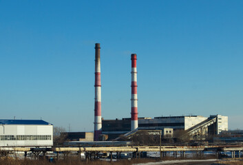 High red and white striped tube lets out white smoke into clear blue sky. Small cloud above pipe on background of electric pole and power wires. Thermal power plant close-up with copy space.