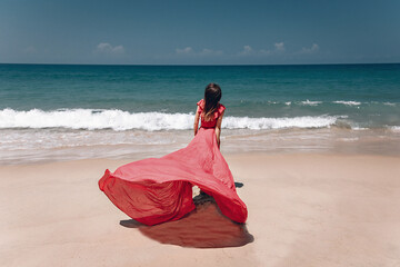 Stunning girl in the exotic sandy beach by the sea. The lady wears light red dress that flows in the wind; fashion concept.