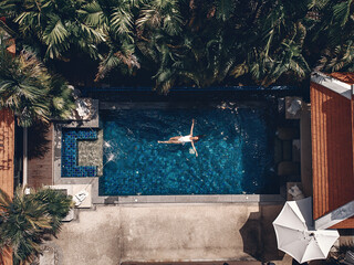 Fototapeta na wymiar Aerial view of the territory of the Thai hotel: tiled terracotta roofs, palm trees, the pool with clear blue water and a young woman swimming there; drone.