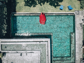 Aerial view of the stunning girl in the pool be the hotel. The young lady wears red dress that flows in the water; fashion concept.