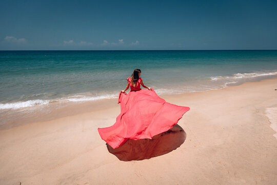 Gorgeous girl in the sunny exotic beach by the ocean. The young woman wears amazing red dress aflutter in the light breeze, backside view; vogue concept.