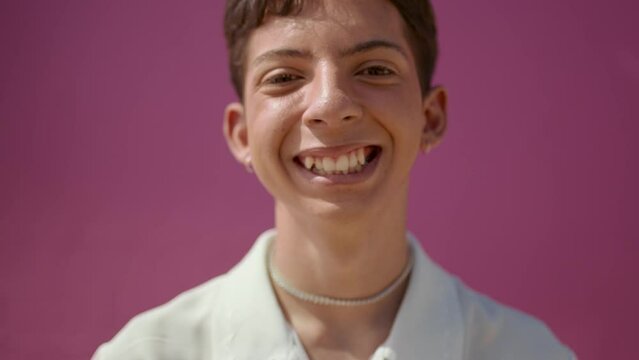 Closeup of a cheerful gay person. Gender queer looking at camera and smiling on pink background.