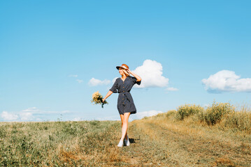 Pretty young woman posing in meadow in summer dress, straw hat and with bouquet of wildflowers on sunny day outdoors. Cute smiling caucasian girl standing on field and looking away, lifestyle