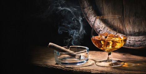 Glass of whiskey with smoking cigar and ice cubes in front of old barrel - obrazy, fototapety, plakaty