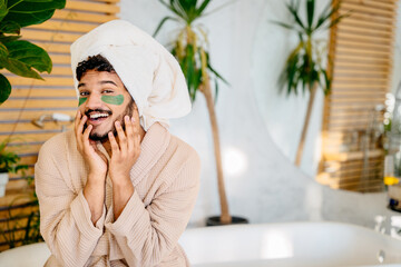 Funny bearded asian man with a green cosmetic pathes under eyes on his face in bathrobe and a towel on his head makes spa treatments on his face resting at home.