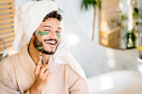 Metrosexual guy with green eye patches using roller for face skincare treatment for glowing skin, have spa day on weekend at home.