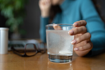 Sick woman sitting at office desk using cold medications to relieve flu symptoms at workplace, selective focus. Closeup of female hand holding glass with effervescent tablet from headache in water