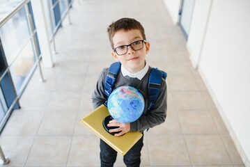 Happy smiling boy in glasses is going to school for the first time. Child with school bag and book in his hand. Kid indoors of the class room . Back to school.