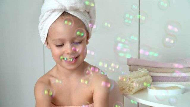 Portrait of little girl with wrapped towel on head laughs and plays soap bubbles in bathroom. Happy kid after bathing. Baby care. Soap and shampoo concept