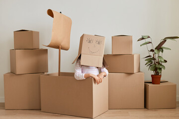 Indoor shot of unknown girl sitting surrounded with packages with personal pile, making head from cardboard box with drown smiling face, relocating to a new apartment.