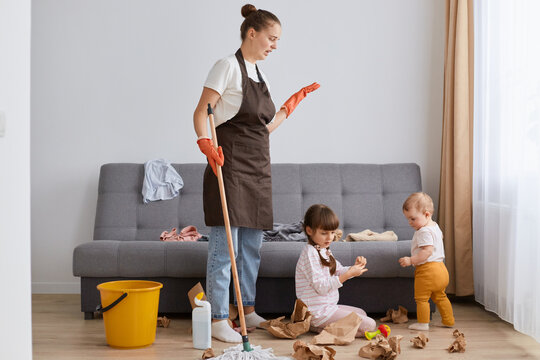 Shocked woman wearing white t shirt, brown apron and jeans, cleaning house, posing with her daughters, washing floor, upset that the kids have done a mess.