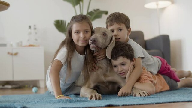 Happy Kids Lying On Floor And Hugging Big Lovely Dog At Home. Front View Of Cheerful Sister And Brothers Having Fun, Playing With Pet, Looking At Camera, Kissing Best Friend. Family, Love Concept