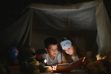 Obraz na płótnie Canvas reading and family games in children's tent. boy and girl with book and flashlight before going to bed