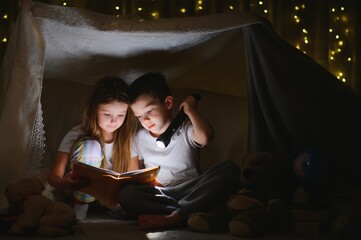 Little kids involving in reading amazing book. They lying in nice toy tent in playroom. Boy holding flashlight in hand