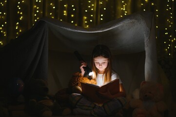 Obraz na płótnie Canvas happy child girl laughing and reading book in dark in a tent at home