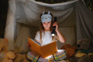 child girl reading with book and flashlight and teddy bear in tent. before going to bed