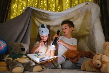 Two children with flashlight read a book under a blanket as a tent - obrazy, fototapety, plakaty