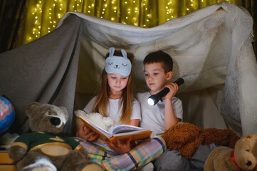 Fototapeta na wymiar Siblings sit in a hut of chairs and blankets. Brother and sister reading book with flashlight at home