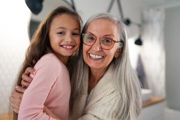 Senior grandmother and granddaughter standing indoors in bathroom, hugging and looking at camera.