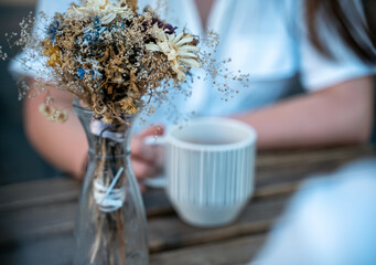Glass vase with dried flowers, beautiful bouquet. Behind the background a woman drinks tea from a mug