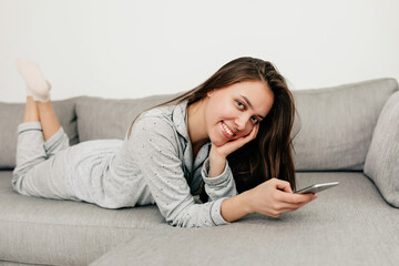 lovely pretty girl with long dark hair is wearing home suit lying on sofa and smiling at camera with smartphone. Indoor portrait of happy girl is relaxing at home