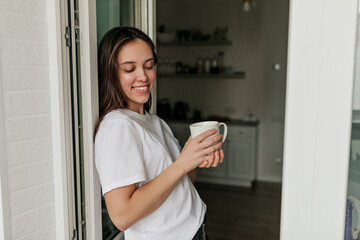 Lovely relaxed girl with straight dark hair and wonderful smile is holding a cup and close eyes leaning against the doorway