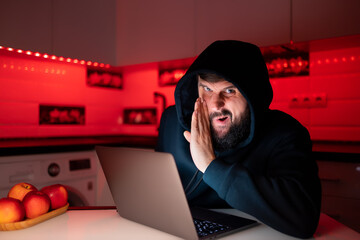a hacker in a black hoodie with a hood on his head is sitting at a laptop and winking angrily