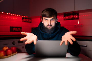 a hacker in a black hoodie is sitting at a laptop, showing his palms forward
