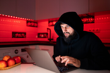 a hacker in a black hoodie is sitting at a laptop hacking the Internet security system - obrazy, fototapety, plakaty
