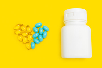 Blue and yellow capsules for treatment in heart shape with white bottle on yellow background, pharmacy and medicine concept, top view