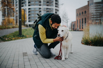 Happy young man squatting and stroking his dog during walk outdoors in city.