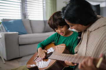Little multiracial boy learning to play the guitar with his mother at home.