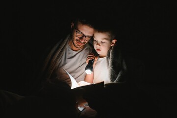 Happy family reading bedtime story under blanket in evening. Father and son spend time together. Father's Day.