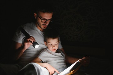 Father and son with flashlight reading book under blanket at home - obrazy, fototapety, plakaty