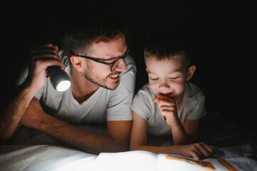 Happy family reading bedtime story under blanket in evening. Father and son spend time together....
