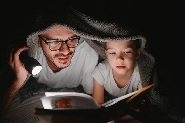 Happy family reading bedtime story under blanket in evening. Father and son spend time together. Father's Day.
