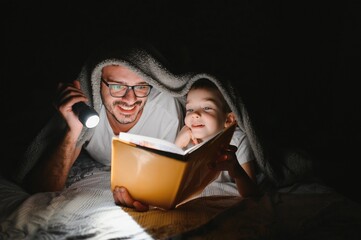 Father and son with flashlight reading book under blanket at home