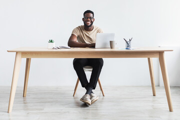 Happy young African American man in casual wear working on laptop at office desk, copy space