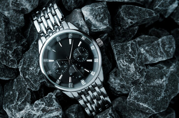 A wristwatch with a black dial and a silver bracelet lies in black stones with white veins close-up.