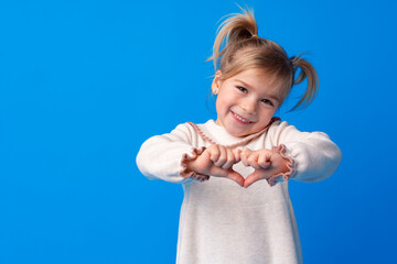 Happy little girl making heart shape by hands on blue background.
