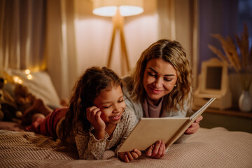 Happy mother with her little daughter lying on bed and reading book in evening at home.