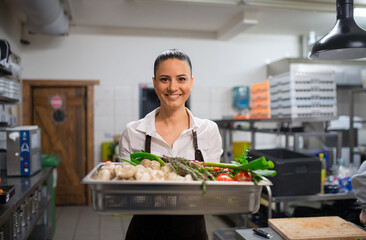 Female chef carrying tray with fresh vegetables and looking at camera in restaurant kitchen.