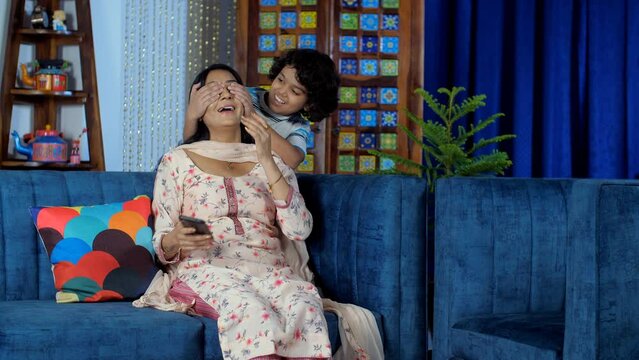An adorable kid in casual clothes hugs his mother from behind - mother-child bonding  love and affection  happy childhood. An attractive female checking her smartphone while sitting on the sofa