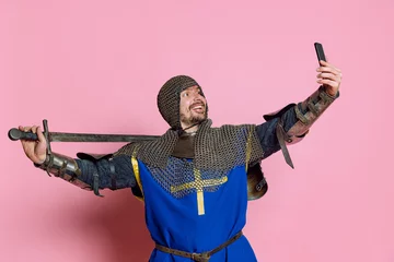 Foto op Plexiglas Half-length portrait of man, medieval warrior or knight in protective chain armor with sword taking selfie on phone isolated over pink background © master1305