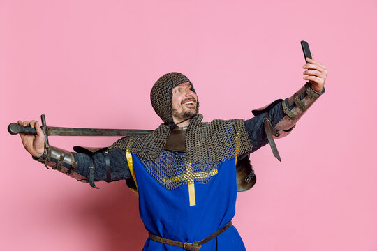Half-length portrait of man, medieval warrior or knight in protective chain armor with sword taking selfie on phone isolated over pink background